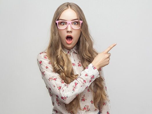 ピンクのふち眼鏡をかけているロングヘアーの女性が、小花柄のシャツを着ていて、左側を指さしている画像