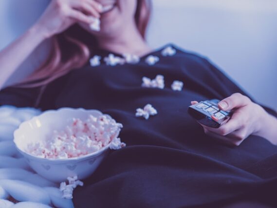女性が寝そべりながらポップコーンを食べてリモコンをもっている画像