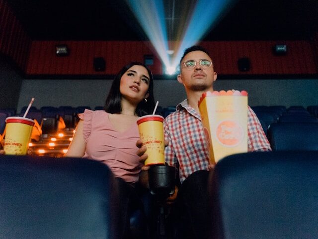 映画館の椅子に男女がジュース都ポップコーンをもって座っている画像