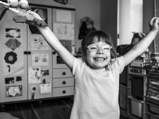 メガネをかけた女の子がおもちゃをもって両手をあげているモノクロ画像