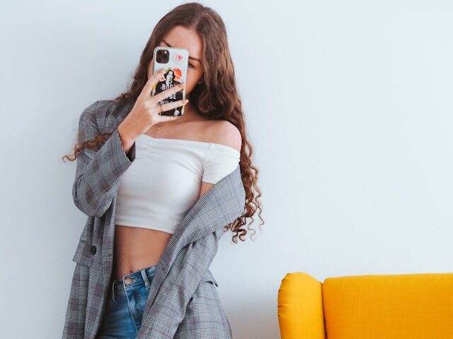 女性が黄色いソファーの隣で携帯を顔の前で持っている画像
